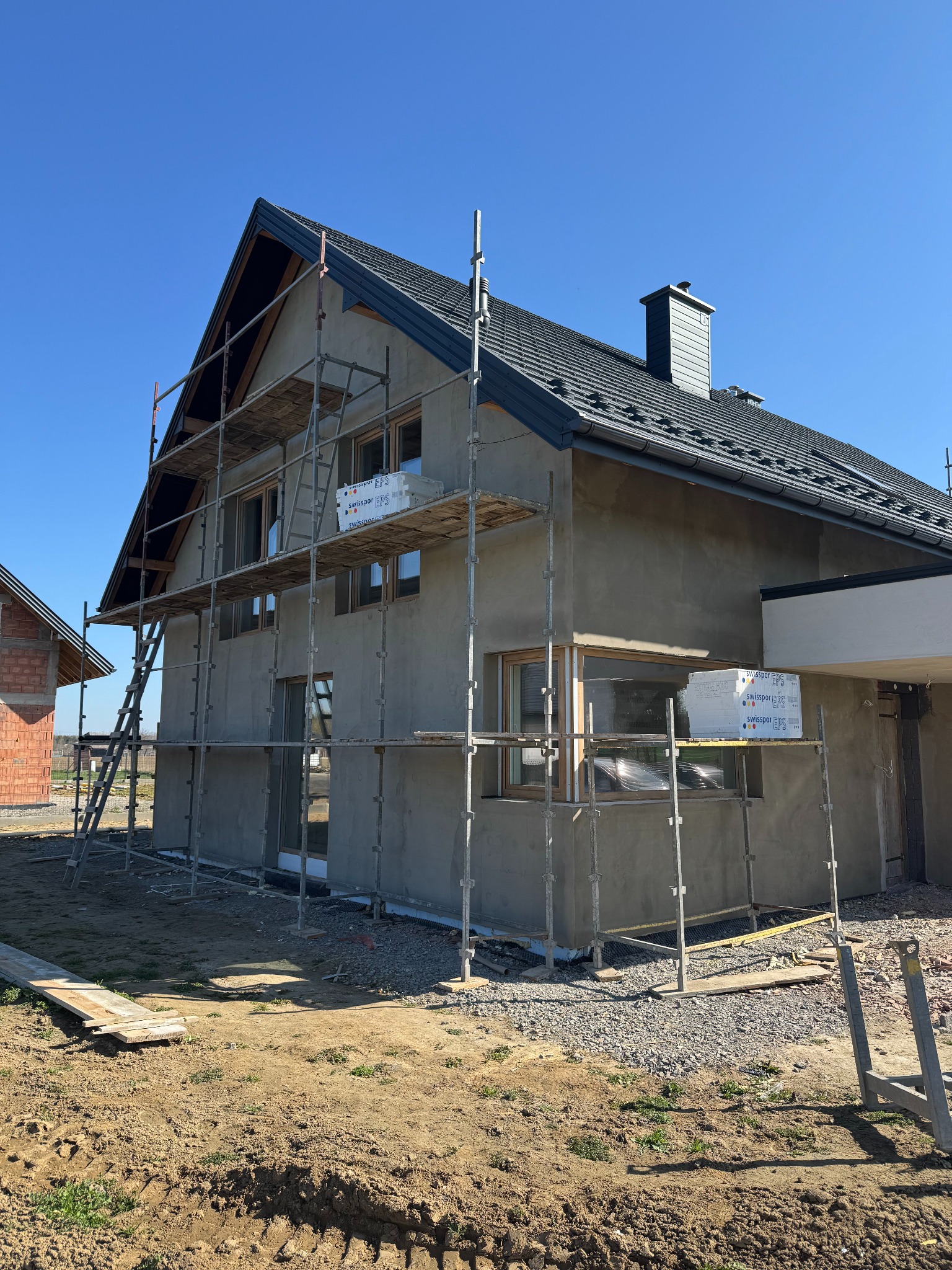 Dom jednorodzinny w trakcie tynkowania, otoczony rusztowaniami. Widoczne okna z drewnianą ramą i dach pokryty dachówką. W tle drugi budynek w stanie surowym.