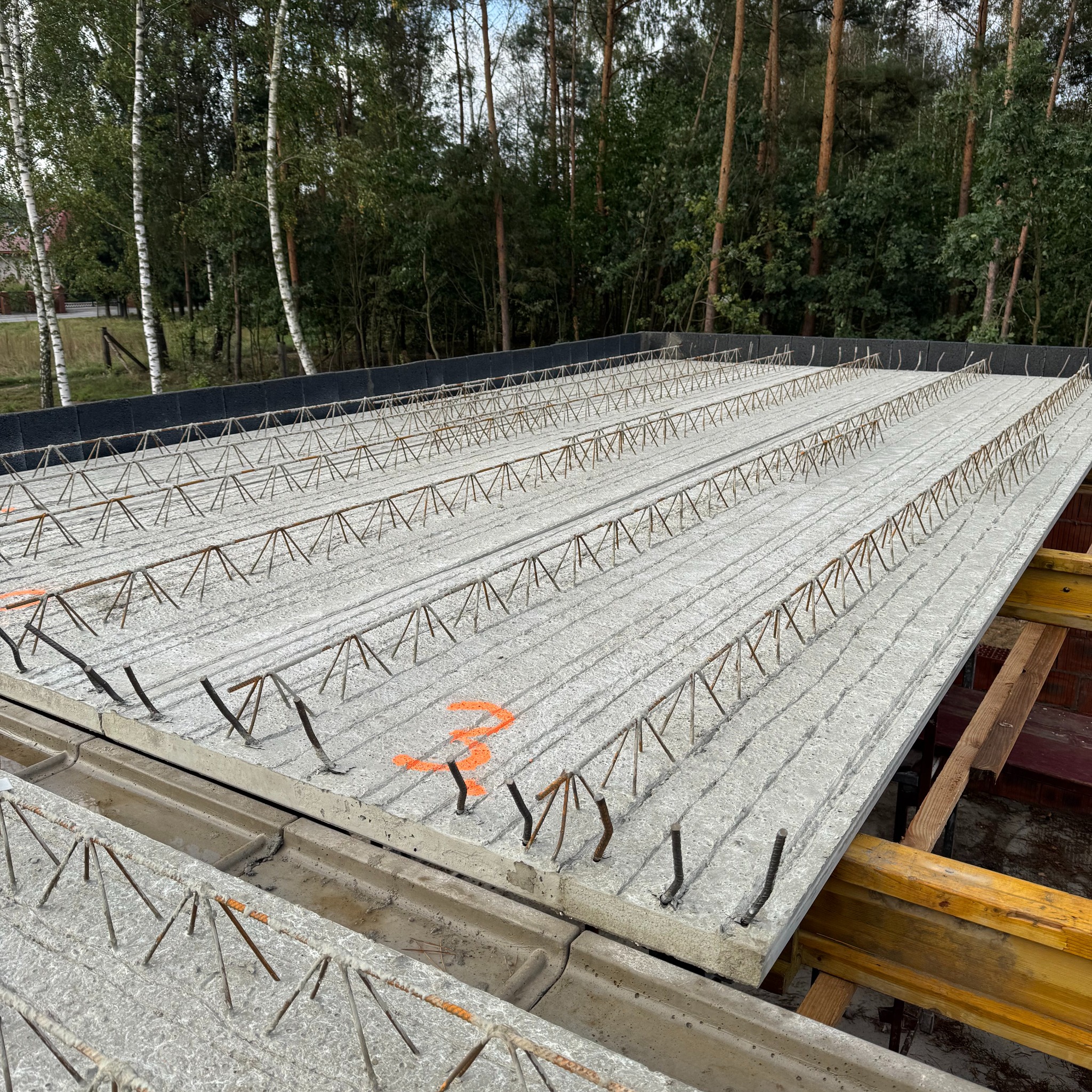 Szara płyta stropowa z wystającymi prętami zbrojeniowymi, ułożona na drewnianych podporach, z widocznym numerem '3' napisanym pomarańczową farbą.