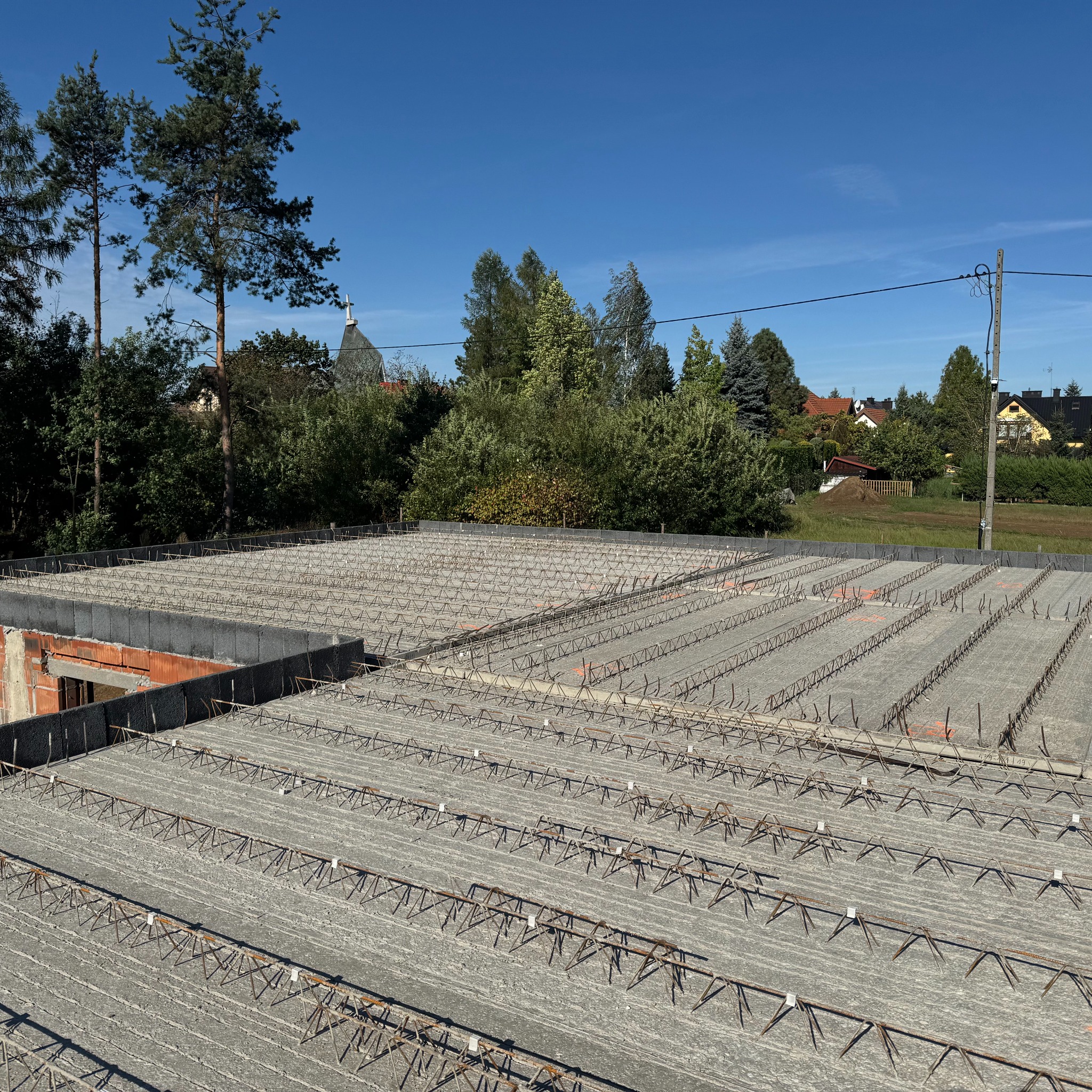 Zbrojenie stropu Teriva na budowie domu jednorodzinnego w słoneczny dzień. Widoczne elementy betonowe i stalowe, w tle zieleń drzew i zabudowa mieszkalna.
