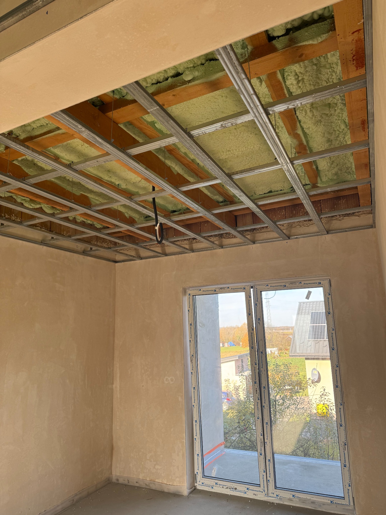 Montaż sufitu podwieszanego z izolacją w nowym budynku. Widoczne profile metalowe, izolacja z wełny mineralnej i fragment okna. Prace wykończeniowe w surowym wnętrzu.