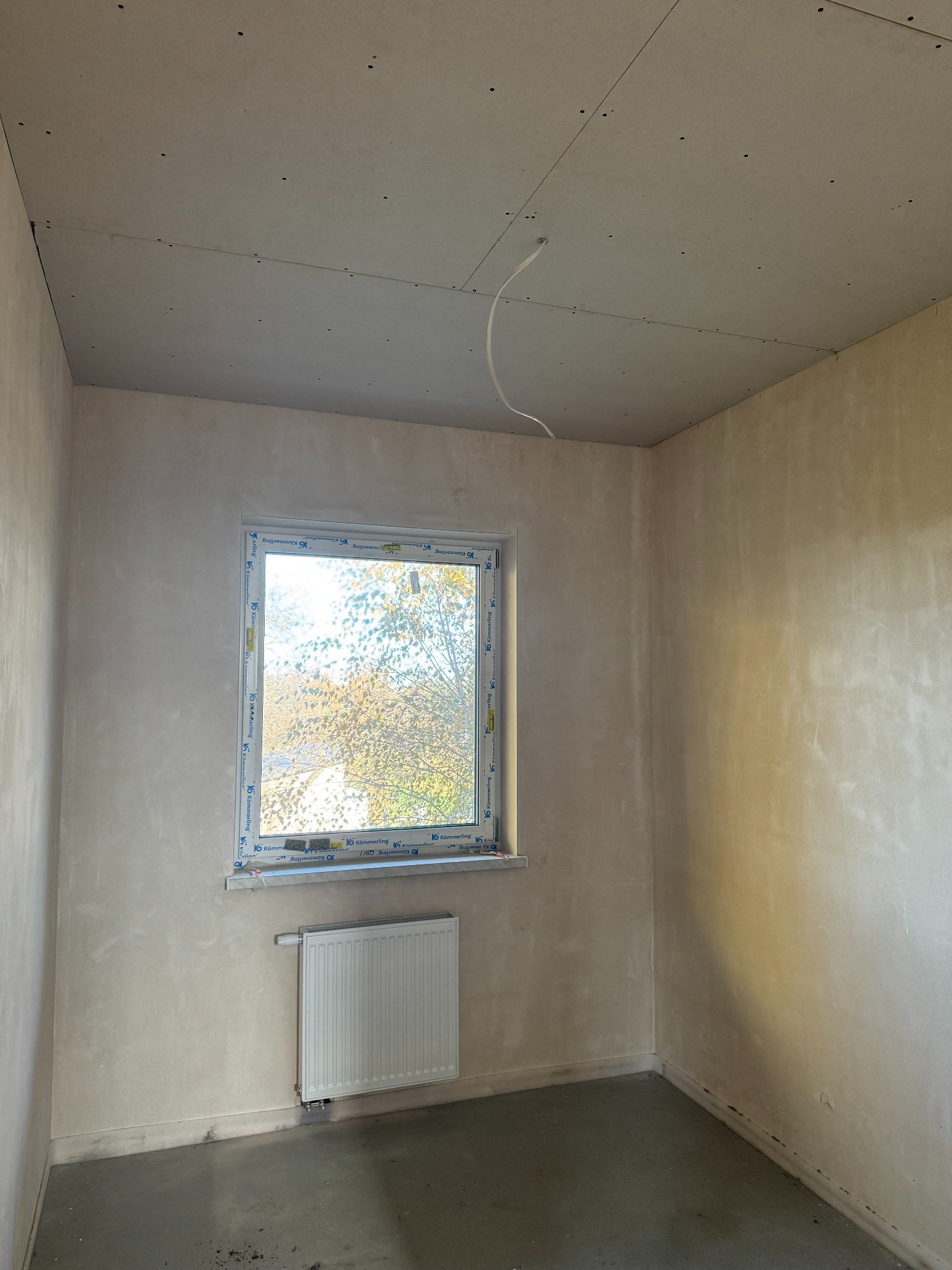 Pokój po tynkowaniu maszynowym: ściany gładkie, okno z widokiem na drzewa, sufit z płyt kartonowo-gipsowych, grzejnik. Widoczne ślady po pracach budowlanych.