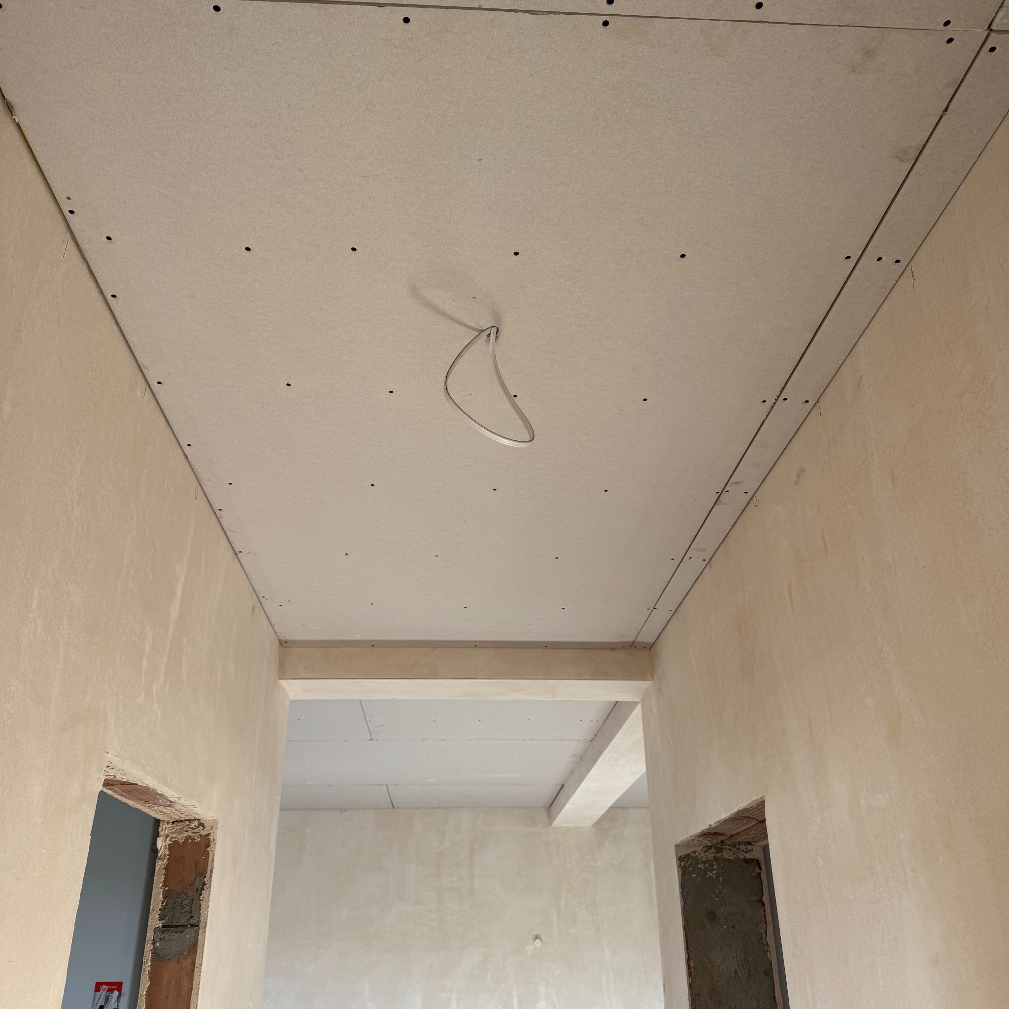 Montaż płyt kartonowo-gipsowych na suficie w surowym wnętrzu. Widoczne profile i przewody elektryczne. Ściany w trakcie tynkowania.