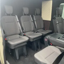 Wnętrze busa z trzema rzędami szarych foteli z pasami bezpieczeństwa, podłoga w kolorze ciemnoszarym, ściany wykończone panelami tapicerowanymi w romby.