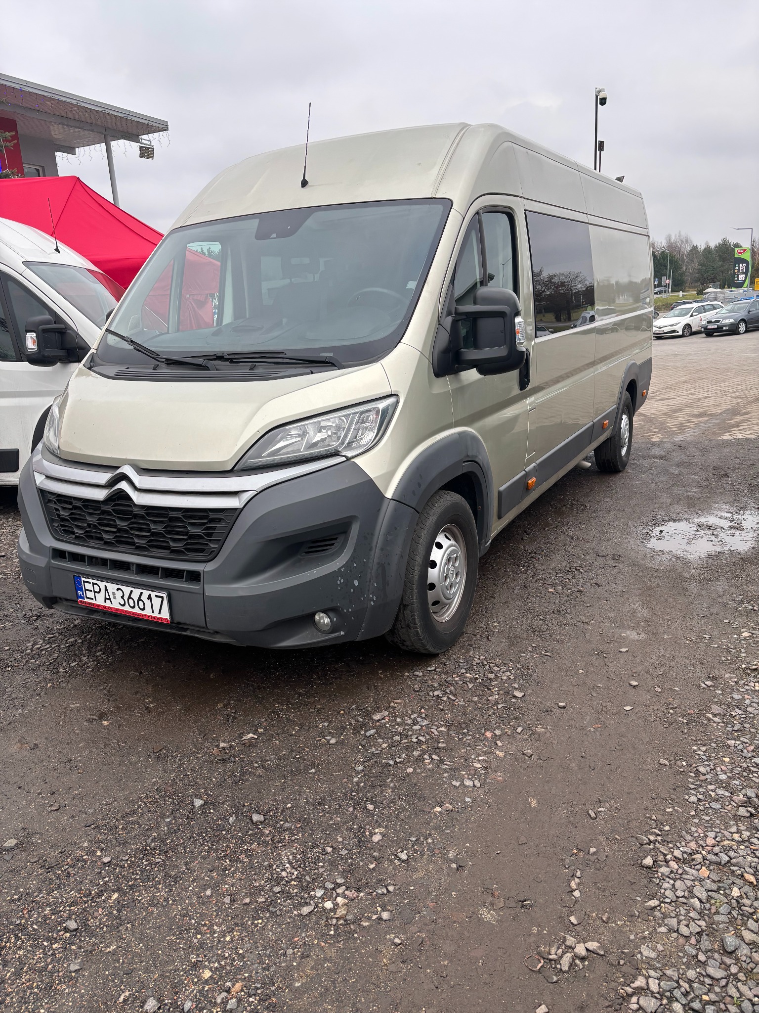 Jasny, beżowy bus Citroen z widoczną tablicą rejestracyjną EPA 36617, zaparkowany na żwirowym parkingu w pochmurny dzień. Z boku widoczny fragment białego busa i czerwony namiot.