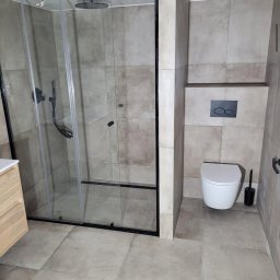 MM CONSULTING MACIEJ MAJCHRZYCKI - Nowoczesna łazienka z kabiną prysznicową typu walk-in z czarnymi ramami, wiszącą toaletą i beżowymi płytkami. Minimalistyczny design, surowy styl.