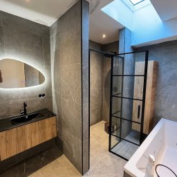 MM CONSULTING MACIEJ MAJCHRZYCKI - Nowoczesna łazienka z szarymi płytkami, podświetlanym lustrem o nieregularnym kształcie i kabiną prysznicową ze szklaną ścianką. Widoczne drewniane szafki i okno dachowe.