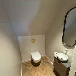 MM CONSULTING MACIEJ MAJCHRZYCKI - Nowoczesna toaleta w skosach: biała ceramika, drewniana podłoga w jodełkę, owalne lustro i umywalka nablatowa. Minimalistyczny design w jasnej tonacji.