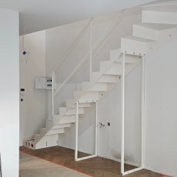 PRO GRAPHIC Jacek Siemiec - Białe schody metalowe z balustradą, wsparte na metalowych nogach, w jasnym wnętrzu w trakcie wykończenia. Widoczna drabina i instalacje.