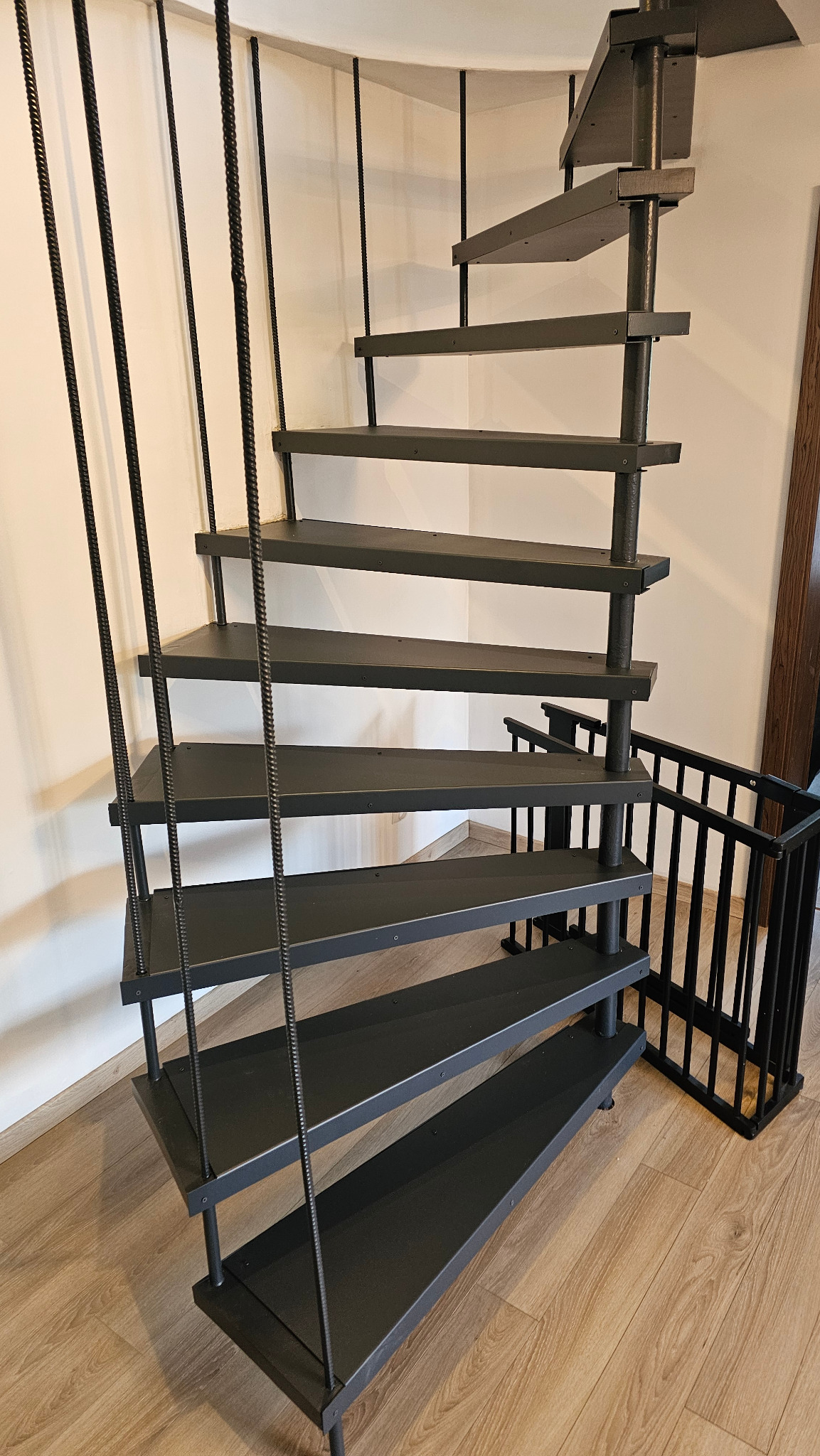 Spiralne schody metalowe w kolorze grafitowym, z stopnicami wykonanymi z blachy stalowej, lakierowane proszkowo. Minimalistyczny design, widok z dołu.