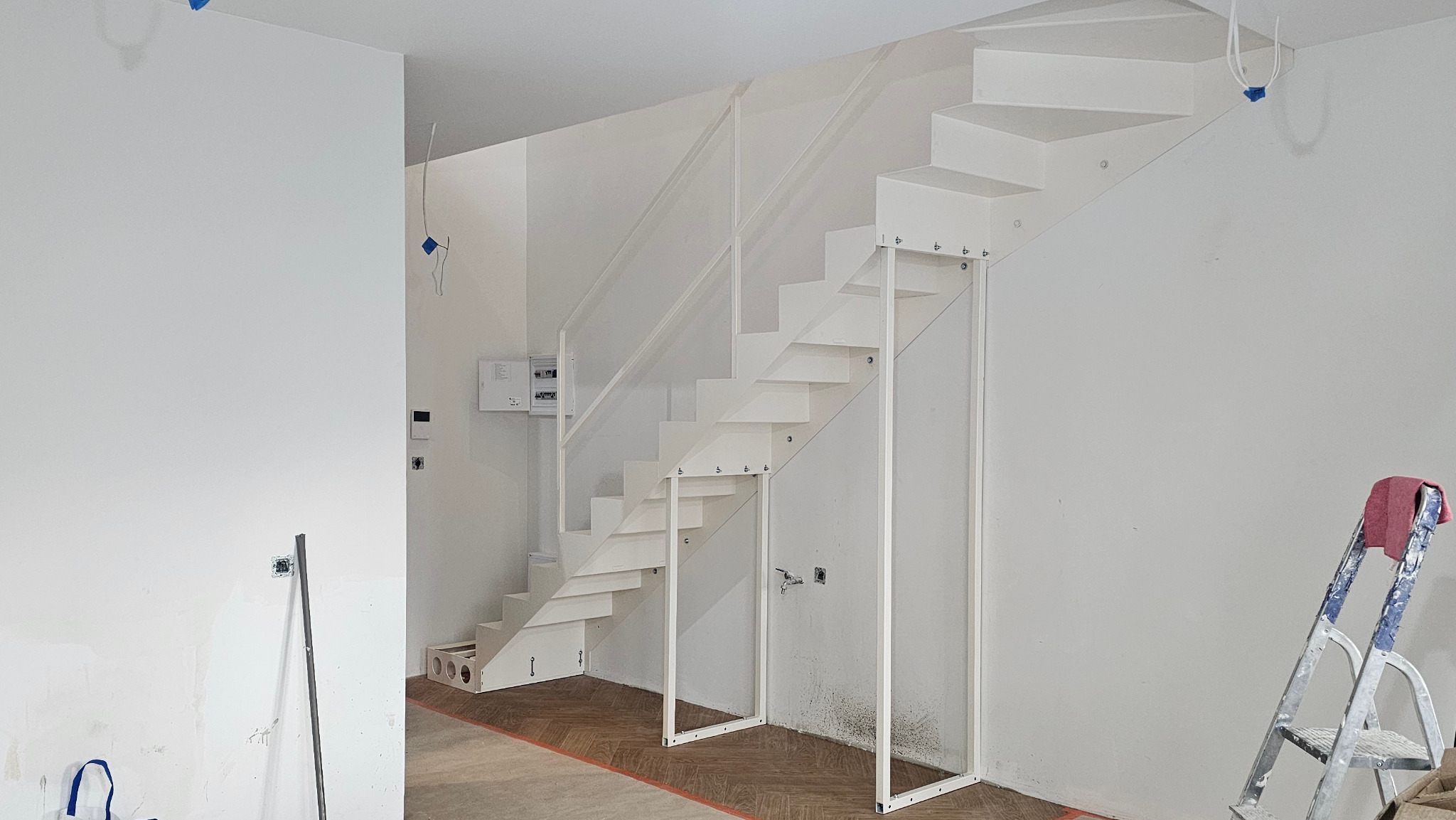 Białe schody metalowe z balustradą, wsparte na metalowych nogach, w jasnym wnętrzu w trakcie wykończenia. Widoczna drabina i instalacje.