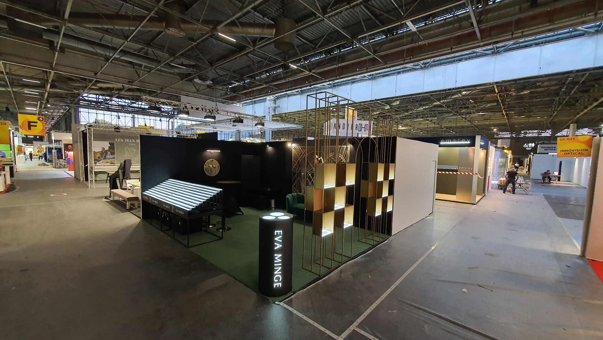 Szeroki kadr na stoisko targowe z podświetlanymi ekspozytorami, ścianką i logotypem 'EVA MINGE', widoczne elementy metalowe i oświetlenie. Hala targowa z innymi stoiskami w tle.