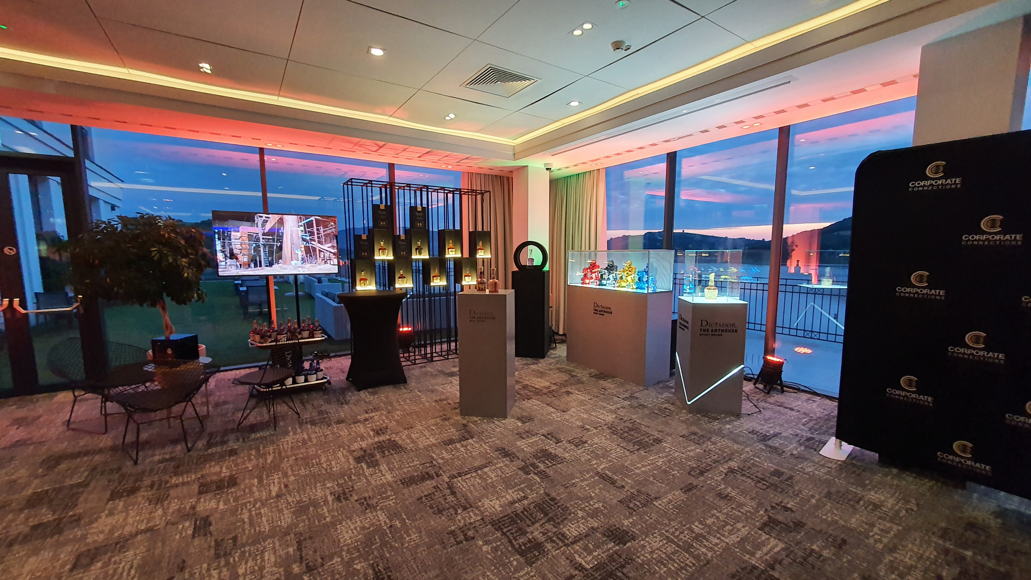 Wnętrze z ekspozycją produktową: metalowy regał z podświetlonymi butelkami, szklane gabloty z kolorowymi figurkami, minimalistyczne postumenty. Za oknami widok na jezioro o zmierzchu.