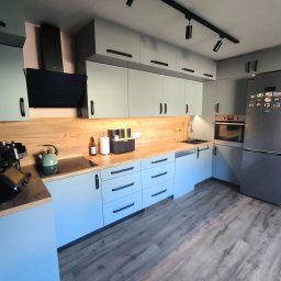 Łukasz Brynkiewicz - Nowoczesna kuchnia z szarymi frontami, drewnianym blatem i czarnymi uchwytami. Widoczne AGD i oświetlenie punktowe. Podłoga wyłożona panelami imitującymi drewno.