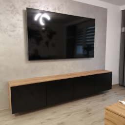Łukasz Brynkiewicz - Czarna szafka RTV z drewnianym blatem, zawieszona pod telewizorem na ścianie o dekoracyjnej fakturze, na tle podłogi z paneli w jasnym odcieniu drewna.