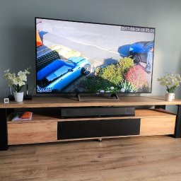 Łukasz Brynkiewicz - Drewniana szafka RTV z czarnymi elementami, na której stoi telewizor z nagraniem monitoringu. Obok głośnik i dekoracje. Podłoga z paneli.