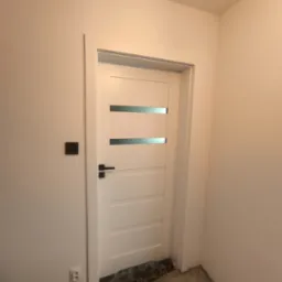 Białe drzwi wewnętrzne z trzema poziomymi przeszkleniami, czarna klamka i kontakt na ścianie. Podłoga w trakcie remontu, widoczne pozostałości po pracach.