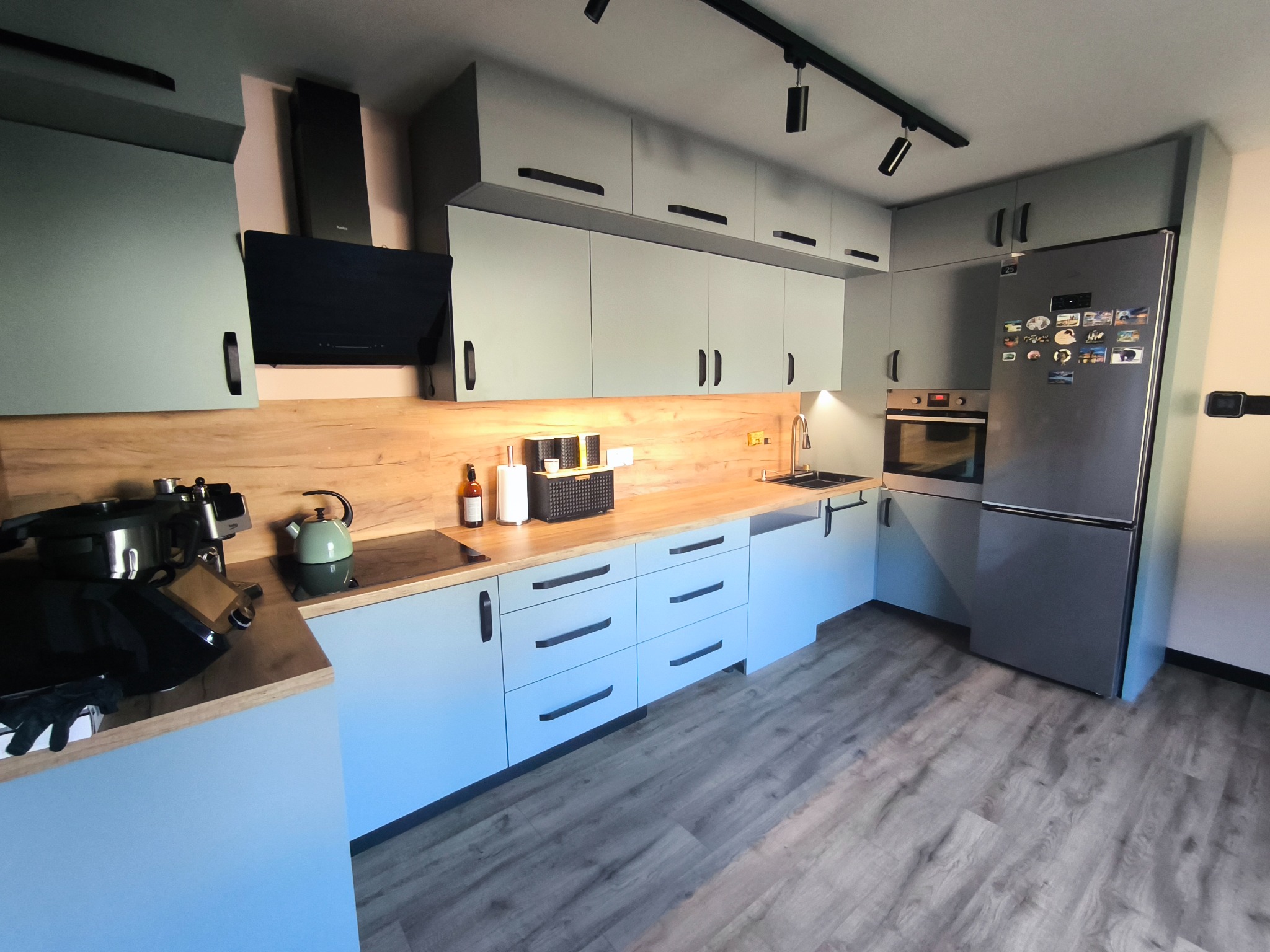 Nowoczesna kuchnia z szarymi frontami, drewnianym blatem i czarnymi uchwytami. Widoczne AGD i oświetlenie punktowe. Podłoga wyłożona panelami imitującymi drewno.