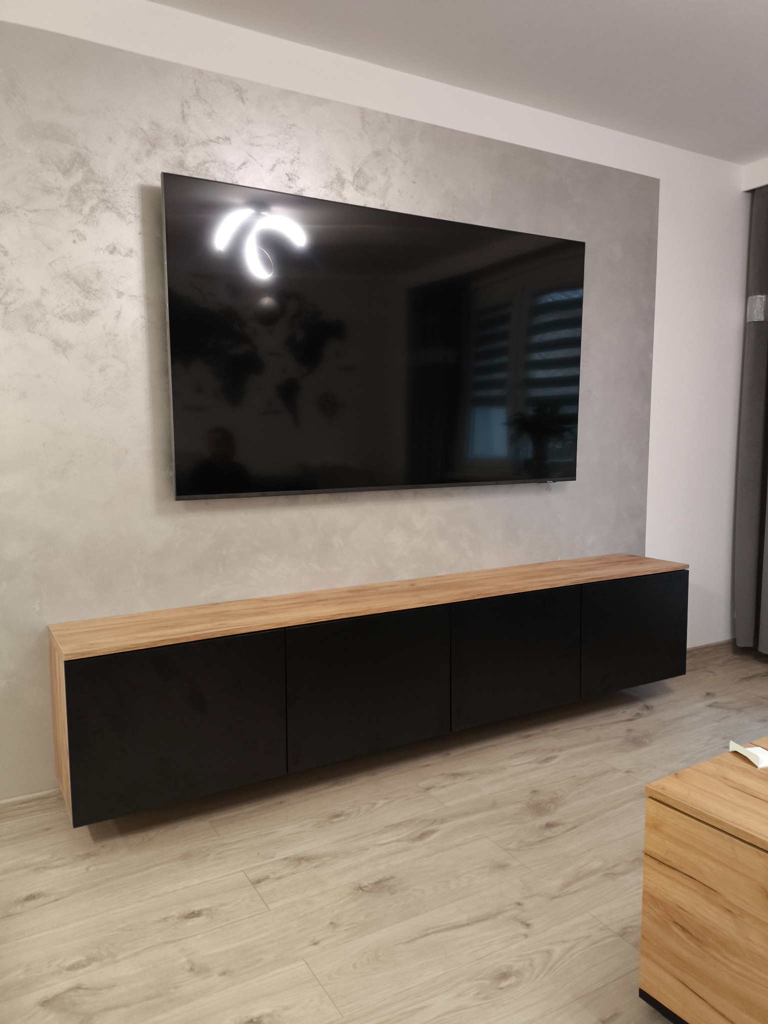 Czarna szafka RTV z drewnianym blatem, zawieszona pod telewizorem na ścianie o dekoracyjnej fakturze, na tle podłogi z paneli w jasnym odcieniu drewna.