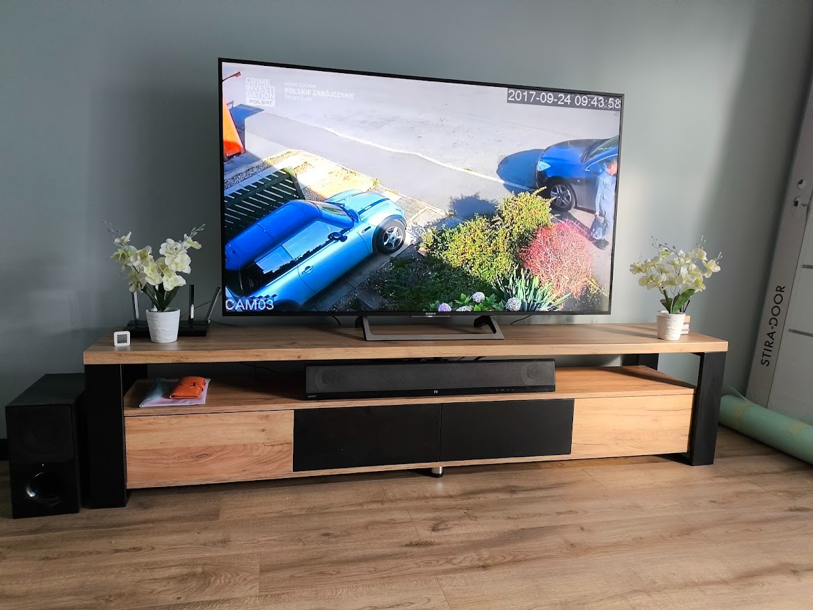 Drewniana szafka RTV z czarnymi elementami, na której stoi telewizor z nagraniem monitoringu. Obok głośnik i dekoracje. Podłoga z paneli.
