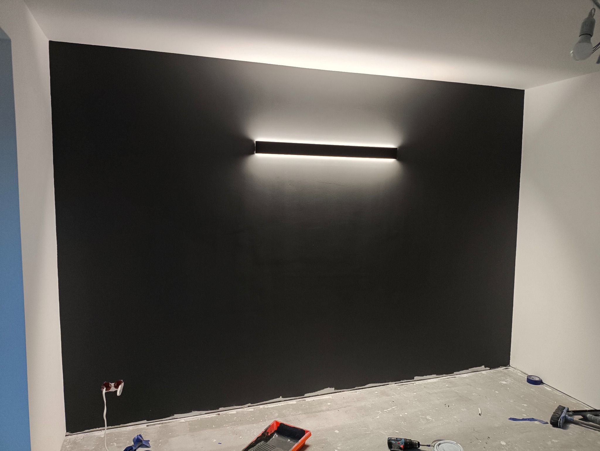 Ściana pomalowana na czarno z podłużną lampą LED. Widoczne narzędzia malarskie i fragmenty pomalowanej podłogi. Wykończone gniazdko elektryczne po lewej.