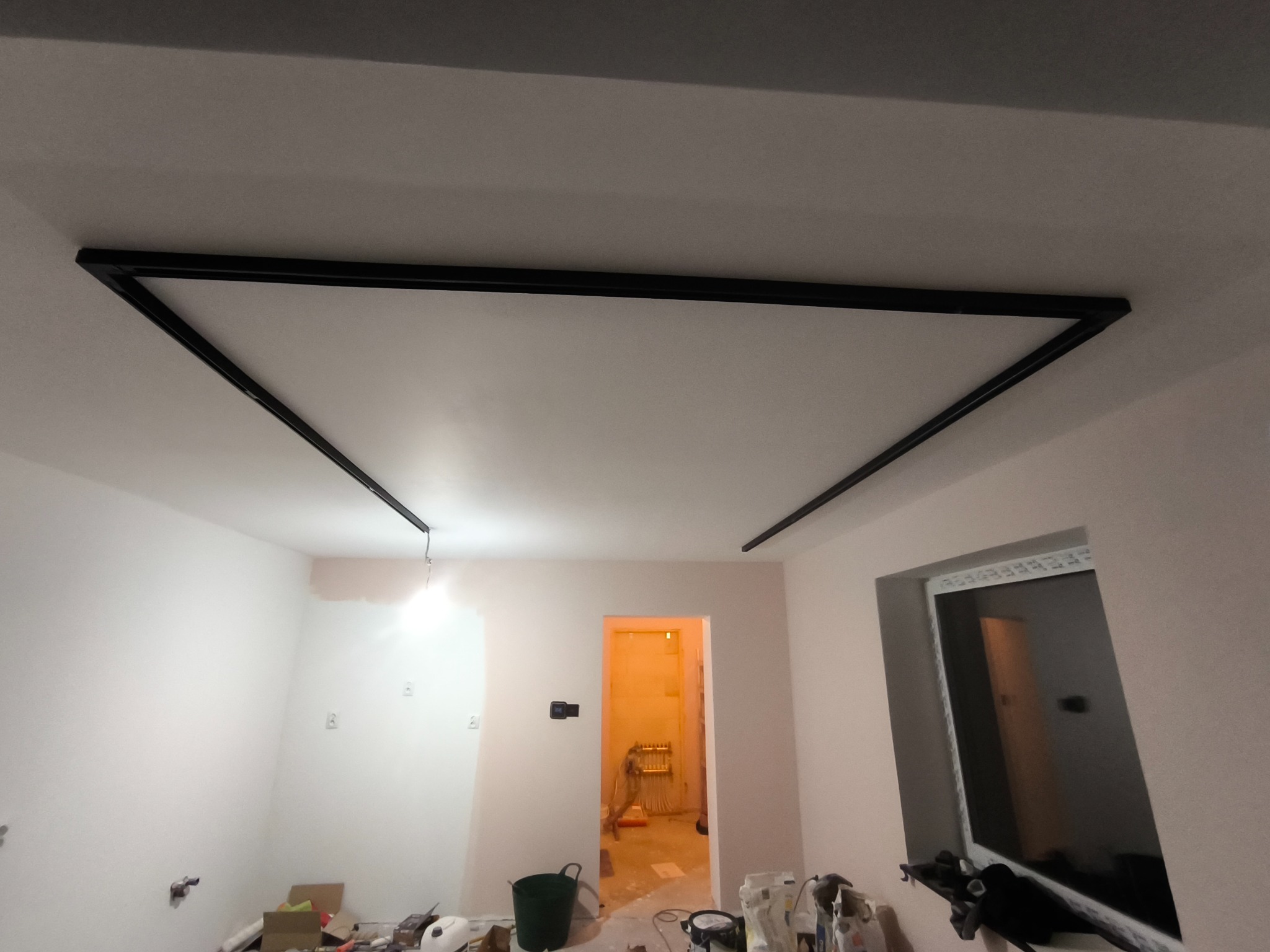 Montaż sufitu podwieszanego z czarnymi profilami w jasnym pokoju. Widoczne narzędzia i materiały budowlane. W tle otwarte drzwi.