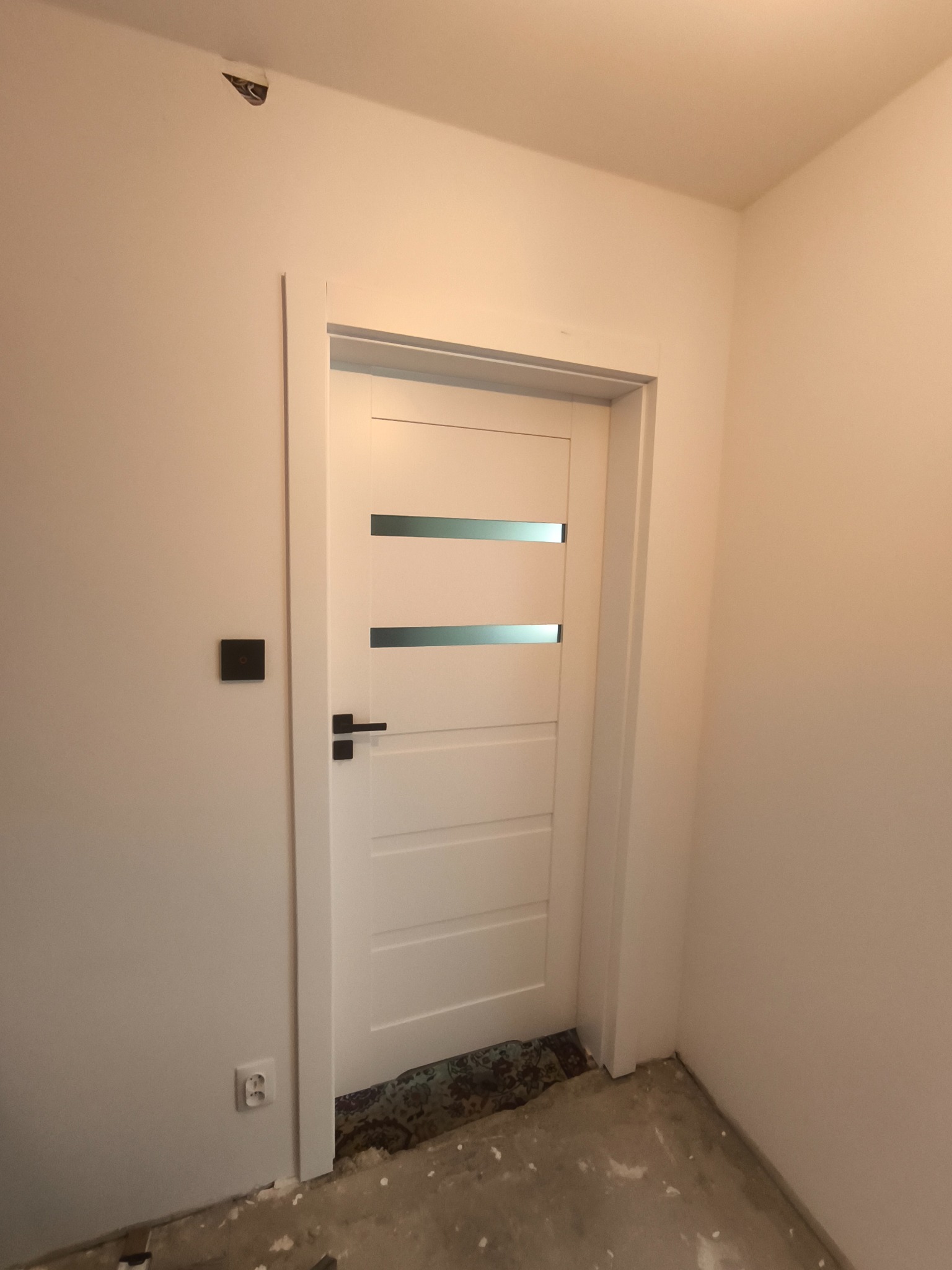 Białe drzwi wewnętrzne z trzema poziomymi przeszkleniami, czarna klamka i kontakt na ścianie. Podłoga w trakcie remontu, widoczne pozostałości po pracach.