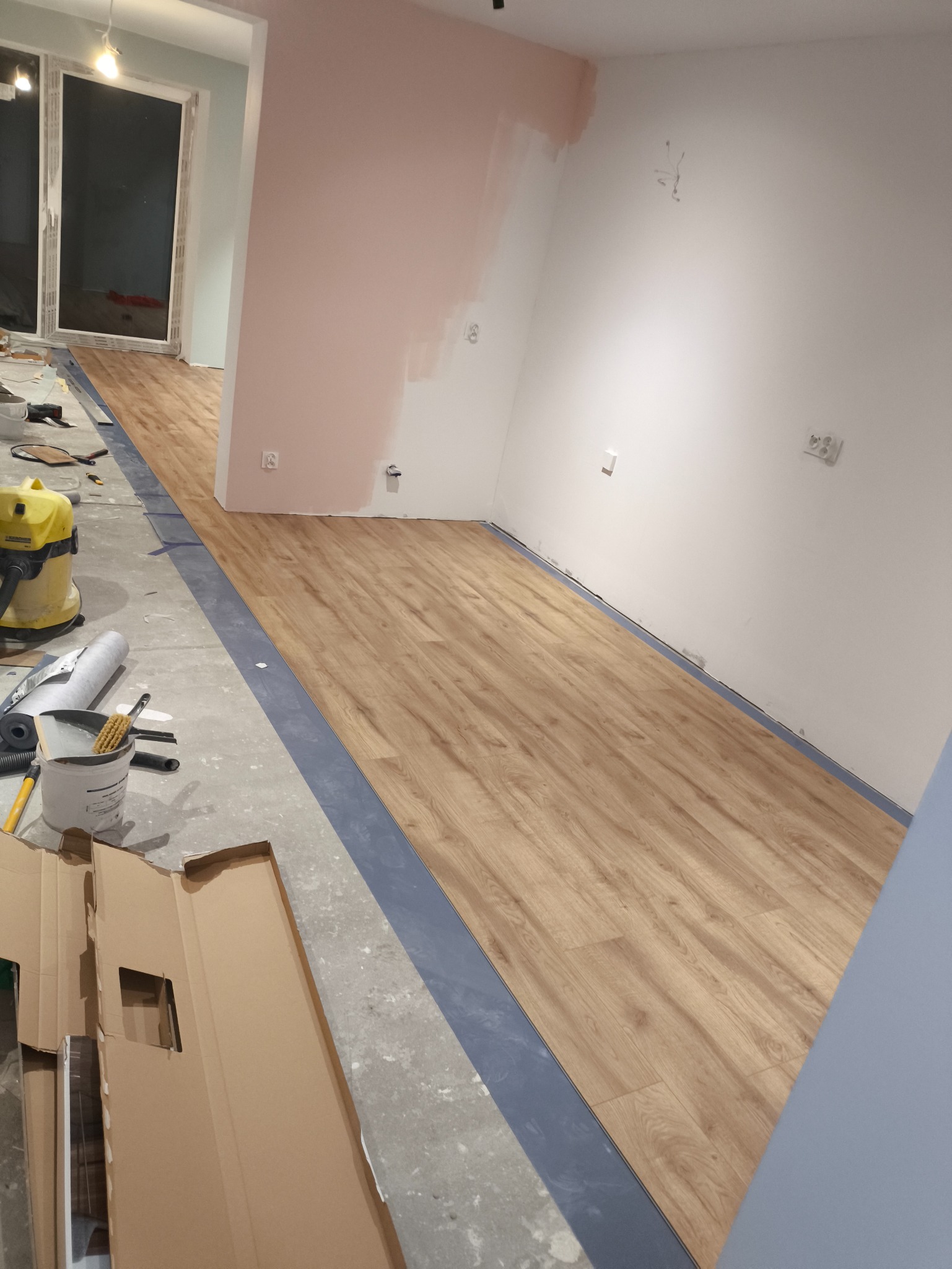 Fragment układania drewnianych paneli podłogowych w pomieszczeniu; widoczne narzędzia i materiały, częściowo ułożona podłoga, przygotowane ściany do malowania.