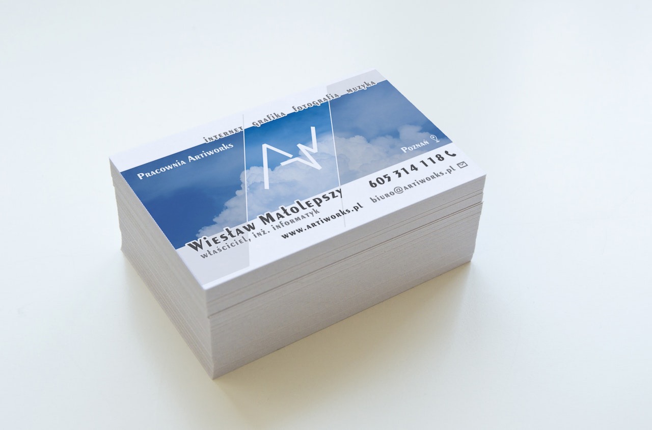 Stos wizytówek Pracowni Artiworks z danymi kontaktowymi: imię, nazwisko, adres e-mail, numer telefonu i adres strony internetowej. W tle niebieskie niebo z chmurami.
