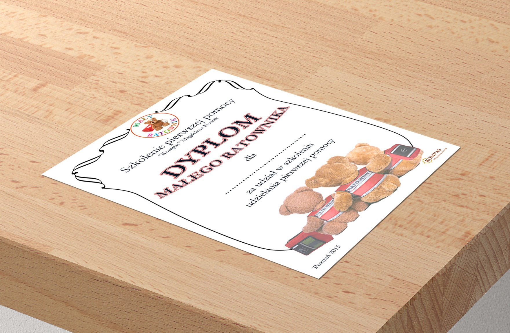 Dyplom Małego Ratownika na drewnianym biurku, z ilustracją pluszowych misiów-ratowników i logo 'Kompas'. Dokument z datą 'Poznań 2015'.