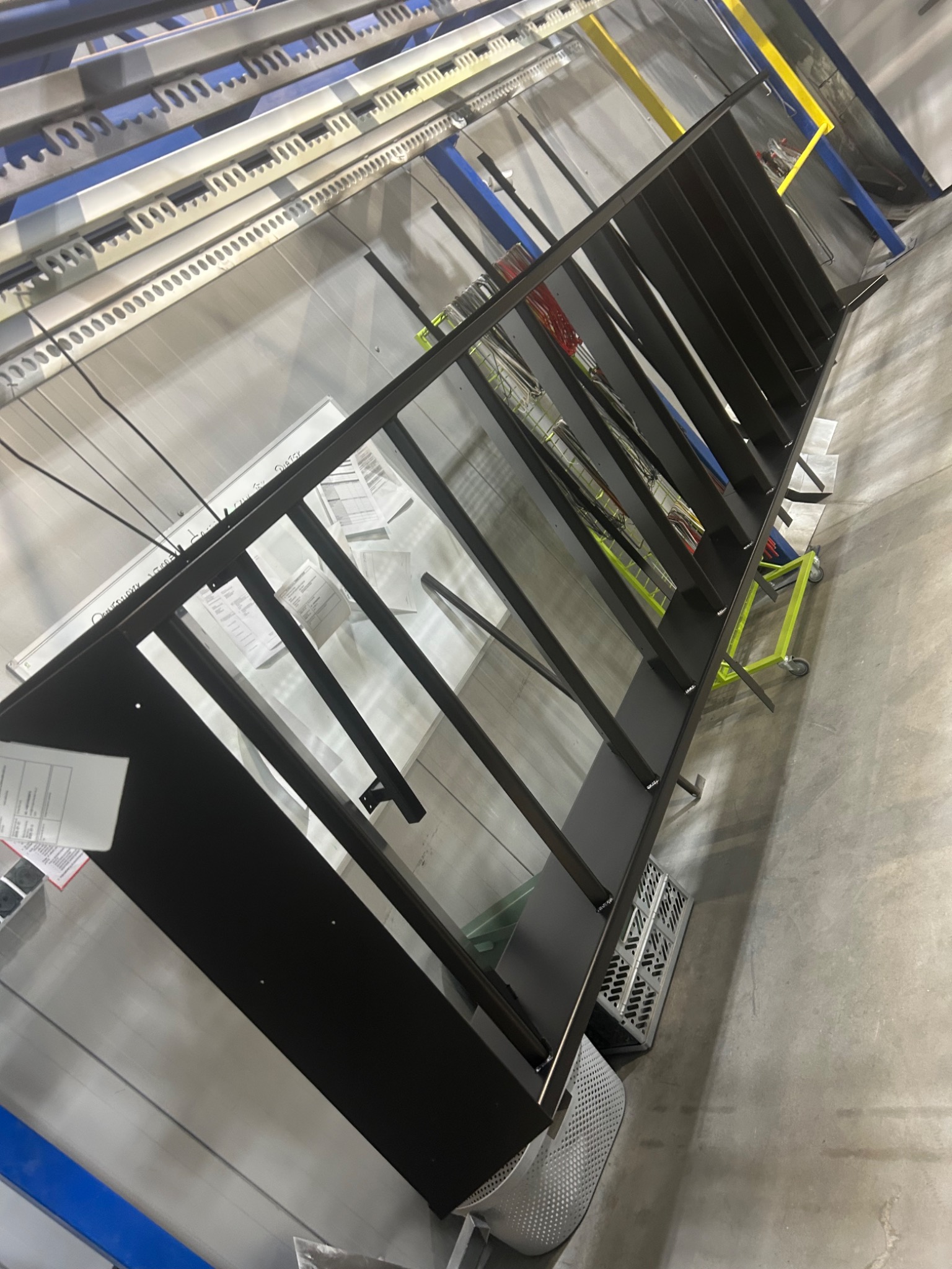 Czarne, metalowe elementy konstrukcyjne w hali produkcyjnej, prawdopodobnie fragment ogrodzenia lub balustrady, widoczne akcesoria montażowe i wózek transportowy.