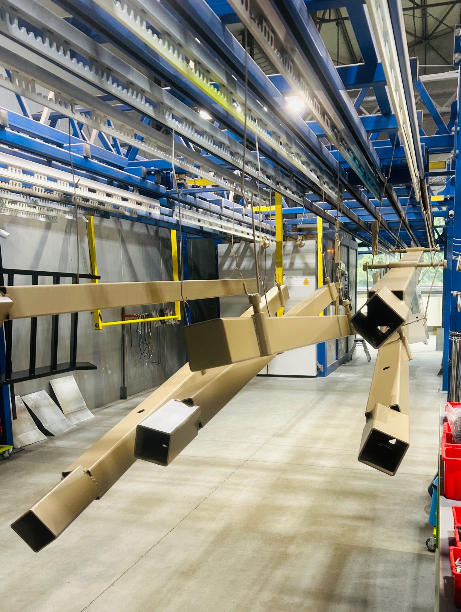 Metalowe elementy konstrukcyjne w kolorze beżowym, zawieszone na systemie transportowym w hali produkcyjnej. Widoczny piec do malowania proszkowego.