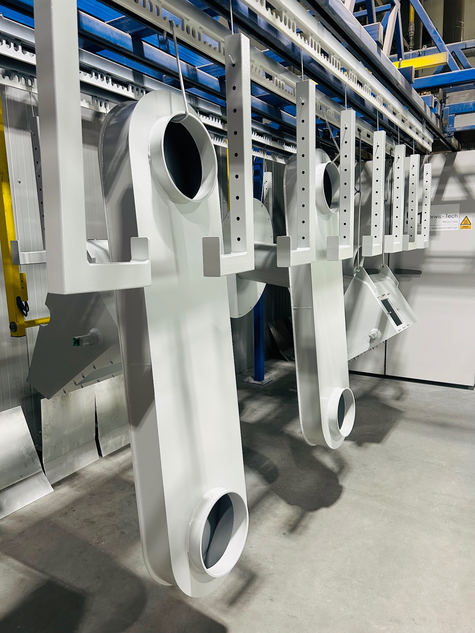 Elementy metalowe w jasnym odcieniu szarości, zawieszone na szynach w hali produkcyjnej. Widoczne otwory i spawane konstrukcje.