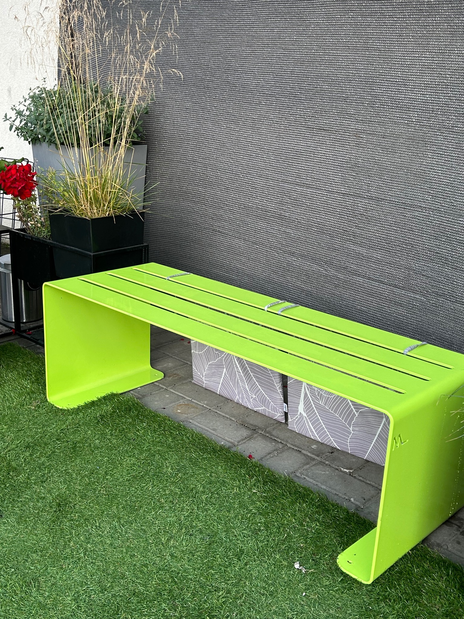 Nowoczesna, limonkowa ławka metalowa z podłużnymi otworami, umieszczona na sztucznej trawie przy szarej, teksturowanej ścianie i donicach z roślinami.