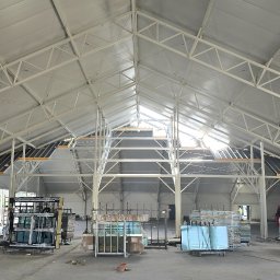 KOPEXPERT Adrian Karcz - Wnętrze budowanej hali z widoczną stalową konstrukcją dachu i przygotowanymi oknami. Na podłodze porozrzucane materiały budowlane, w tle pracownicy budowlani.