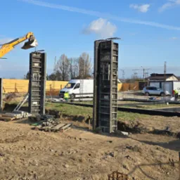 Żółta koparko-ładowarka JCB przy szalunkach kolumn fundamentowych na placu budowy. W tle widoczny biały bus i domy jednorodzinne.
