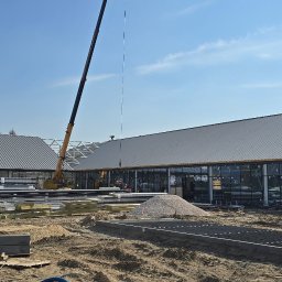 KOPEXPERT Adrian Karcz - Budowa hali z szarej blachy trapezowej, widoczny dźwig i materiały budowlane. W tle błękitne niebo. Prace ziemne w toku, teren przygotowywany pod dalsze etapy budowy.