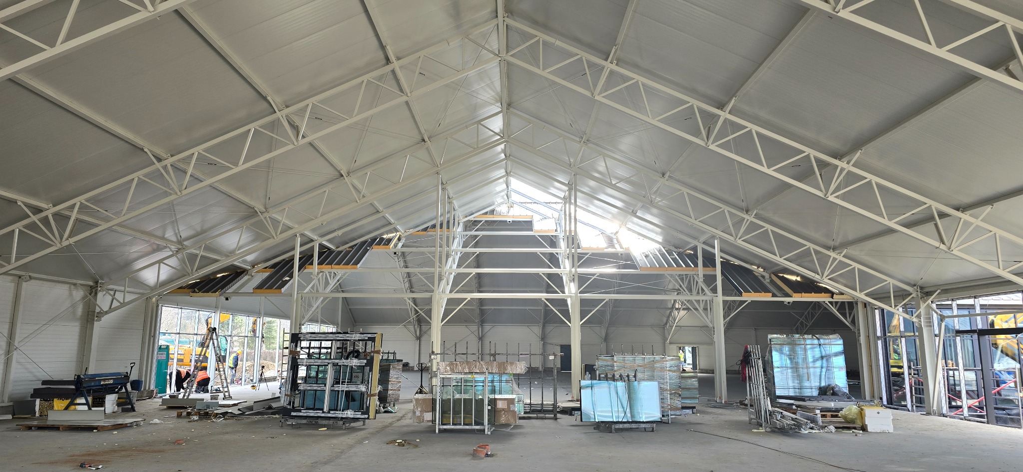 Wnętrze budowanej hali z widoczną stalową konstrukcją dachu i przygotowanymi oknami. Na podłodze porozrzucane materiały budowlane, w tle pracownicy budowlani.