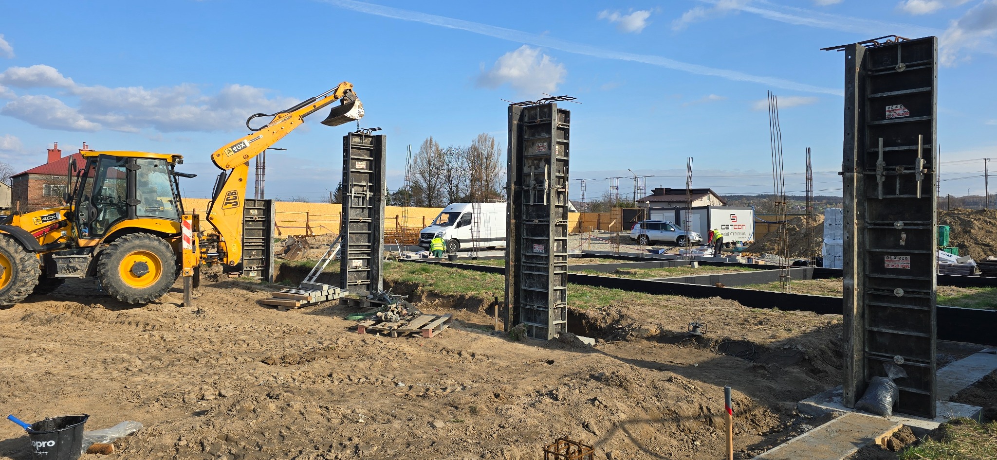 Żółta koparko-ładowarka JCB przy szalunkach kolumn fundamentowych na placu budowy. W tle widoczny biały bus i domy jednorodzinne.