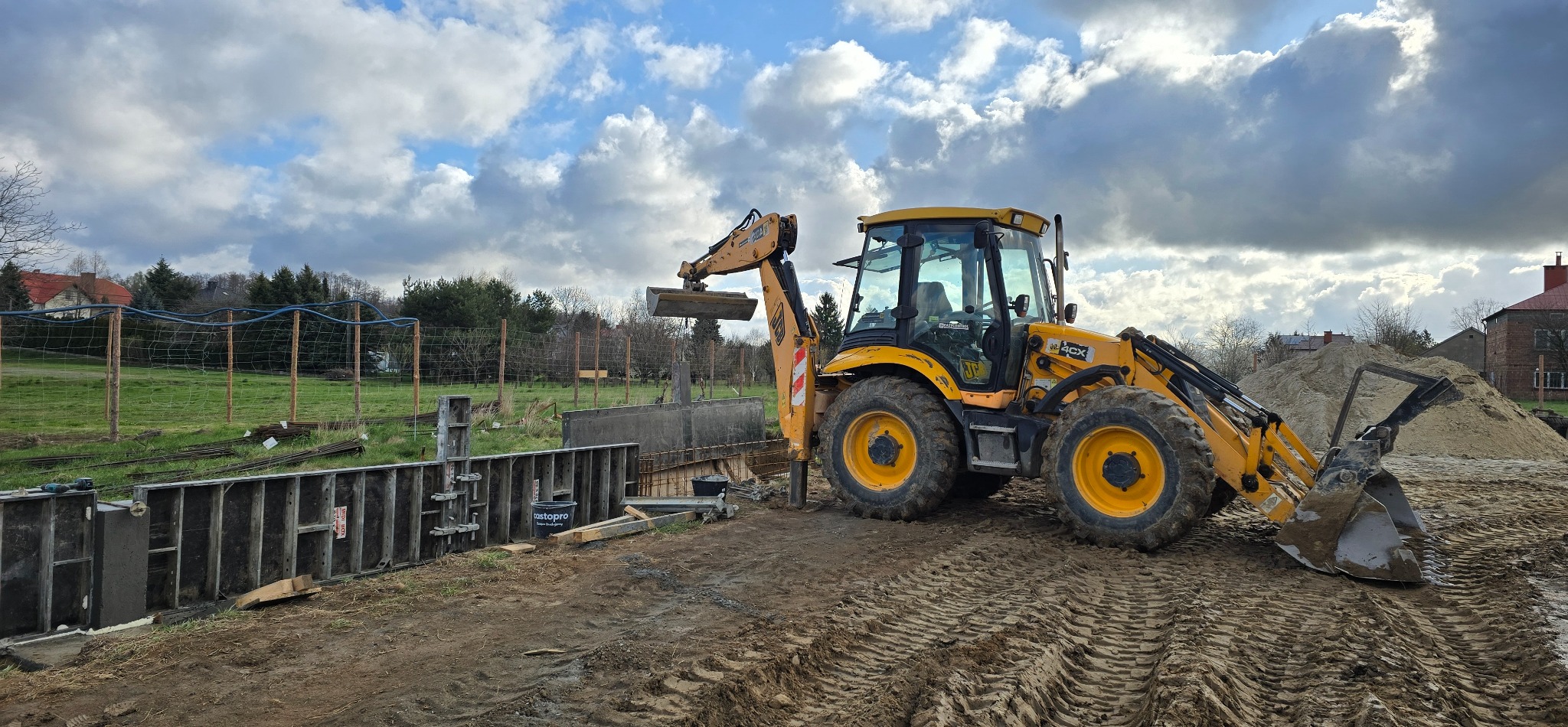 Żółta koparko-ładowarka JCB na placu budowy, obok szalunek fundamentowy. W tle widoczne domy i drzewa pod błękitnym niebem z chmurami.