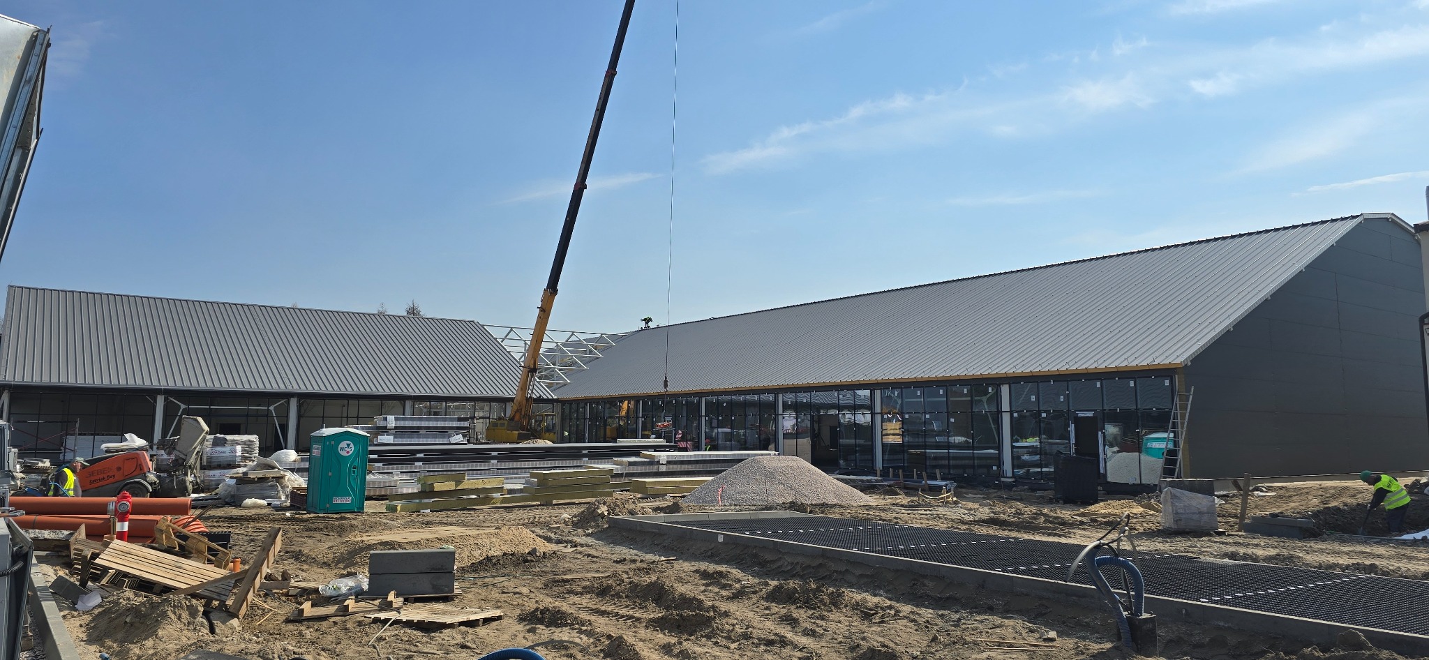 Budowa hali z szarej blachy trapezowej, widoczny dźwig i materiały budowlane. W tle błękitne niebo. Prace ziemne w toku, teren przygotowywany pod dalsze etapy budowy.