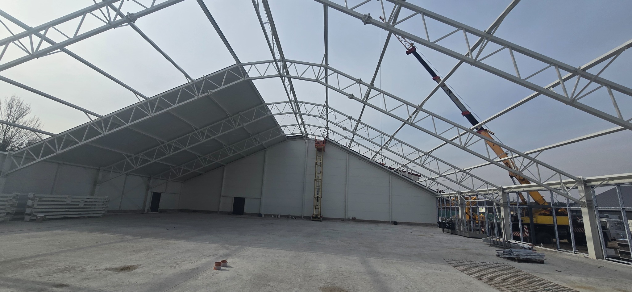Wnętrze hali w budowie z widoczną konstrukcją stalową, częściowo obudowaną płytami warstwowymi i dźwigiem. Budowa hali w trakcie realizacji.
