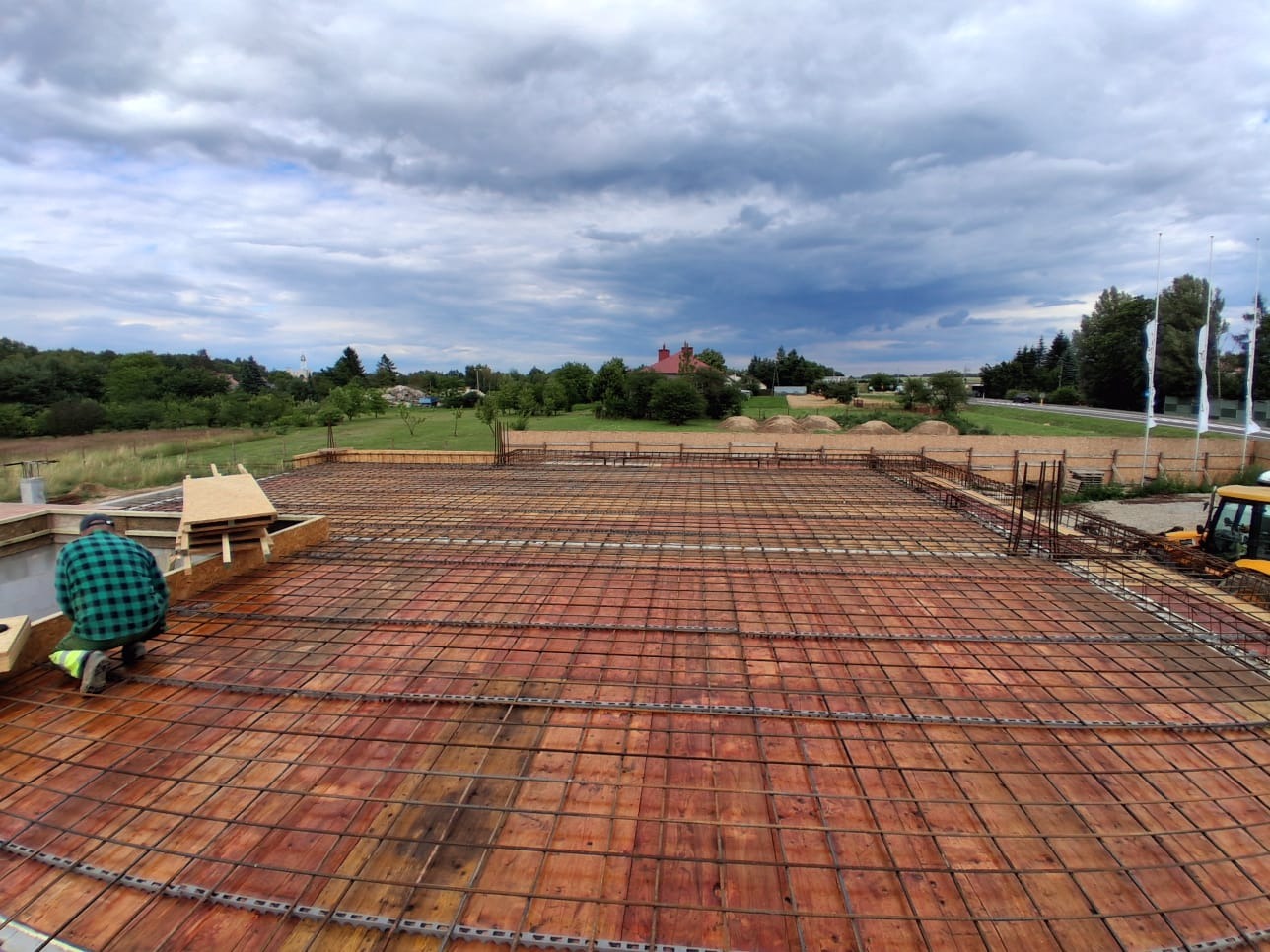 Zbrojenie płyty fundamentowej z drewnianym szalunkiem na tle malowniczego krajobrazu. Widoczny pracownik w zielonej koszuli w kratę i fragment żółtej maszyny budowlanej.