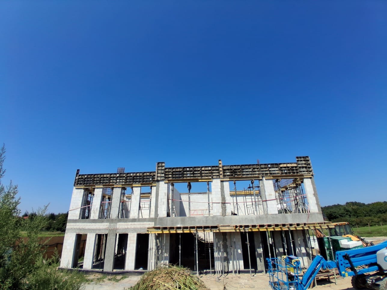 Budowa domu z betonu komórkowego w trakcie realizacji, widoczne szalunki i podnośnik koszowy na tle błękitnego nieba.