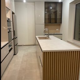 ORZEŁ MEBLE KAMIL ORŁOWSKI - Nowoczesna kuchnia z wyspą: jasne fronty, drewniane lamele na wyspie, stalowe AGD. Minimalistyczny design, neutralna kolorystyka, duże okno z żaluzjami.
