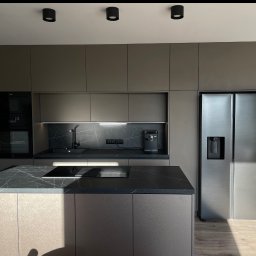 ORZEŁ MEBLE KAMIL ORŁOWSKI - Nowoczesna kuchnia w odcieniach szarości z wyspą i blatem imitującym marmur. Na blacie płyta indukcyjna. W tle zabudowa kuchenna i duża lodówka.