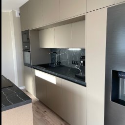 ORZEŁ MEBLE KAMIL ORŁOWSKI - Nowoczesna kuchnia: beżowe szafki, czarny blat z marmurowym wzorem, srebrna lodówka z dystrybutorem wody. Oświetlenie punktowe nad blatem i drewniana podłoga.