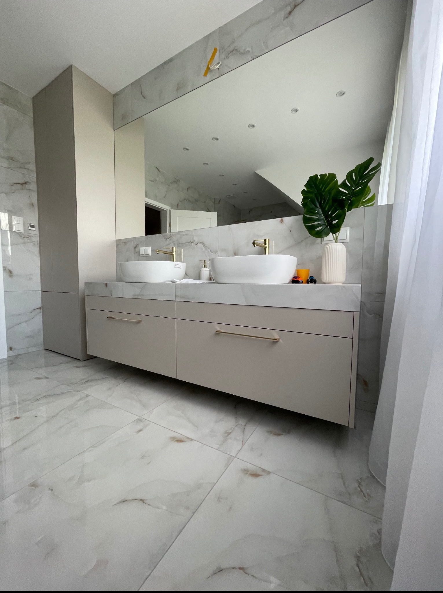 Nowoczesna łazienka z szafką pod umywalki w kolorze beżowym, marmurową podłogą i blatem. Nad blatem duże lustro i dwie białe umywalki.