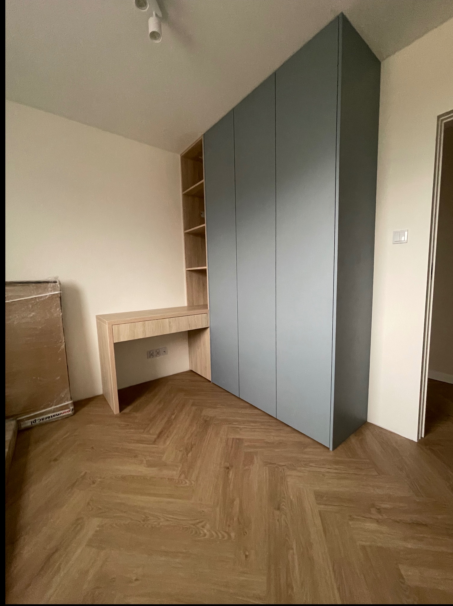 Szara szafa wnękowa z biurkiem z jasnego drewna, parkiet w jodełkę. Widok na fragment pokoju z oświetleniem punktowym. Nowoczesne rozwiązanie do przechowywania i pracy.