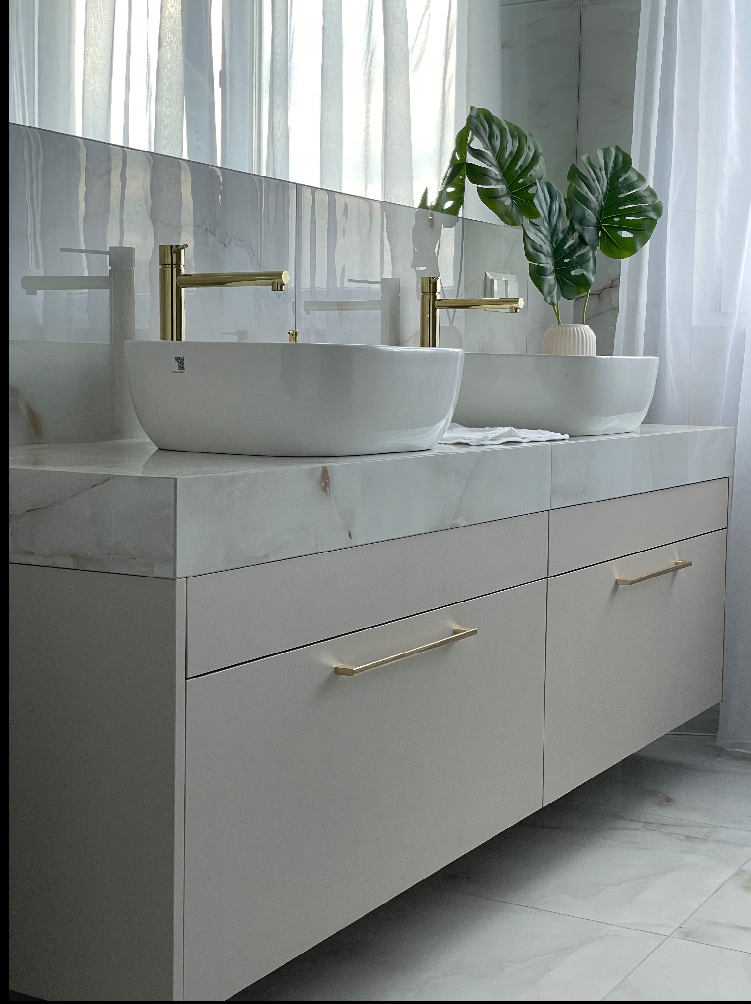 Beżowa szafka łazienkowa z marmurowym blatem, na którym stoją dwie białe umywalki i złote baterie. W tle białe zasłony i płytki imitujące marmur.