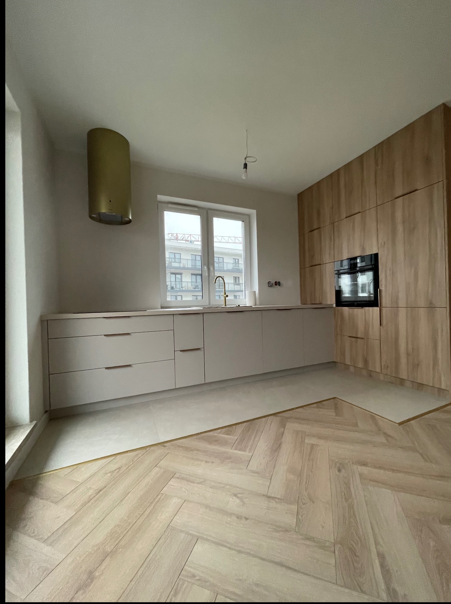 Nowoczesna kuchnia z drewnianymi szafkami i złotym okapem na tle okna z widokiem na budynek. Parkiet ułożony w jodełkę dodaje elegancji.